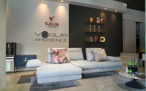 Divano componibile grigio chiaro con cuscini decorativi, logo Calia Italia e "Vogue Xperience" sulla parete.