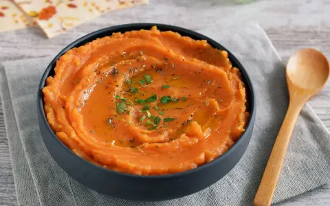 Un tazón de cremoso puré de boniato, adornado con aceite y hierbas, junto a una cuchara de madera.