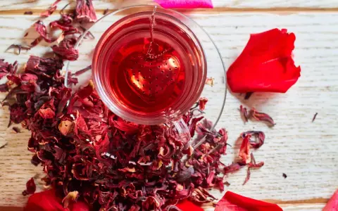 Czerwona, aromatyczna herbata z hibiskusem w szklance z zaparzaczem w kształcie serca, otoczona suszonymi kwiatami i płatkami róż.