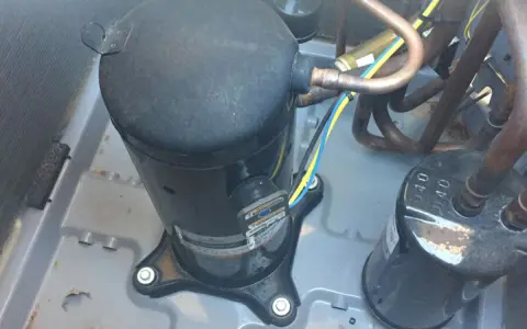Close-up of an AC pump replacement in progress. A large black compressor sits on a gray base, connected to copper tubing and wires.