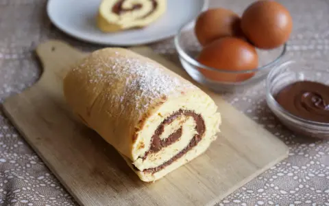 Un delizioso rotolo alla nutella, spolverato di zucchero a velo, pronto per essere gustato. Ingredienti freschi, uova e crema al cioccolato, completano la scena.