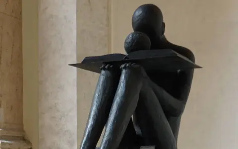 Statua di una figura che legge un libro, con un bambino appoggiato, immersi nella conoscenza a tutto spiano.