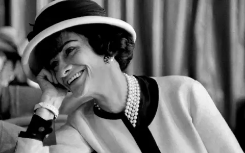 Coco Chanel sorride con un cappello e perle, un'icona di stile che sembra raccontare la storia di quando è nata Coco Chanel.
