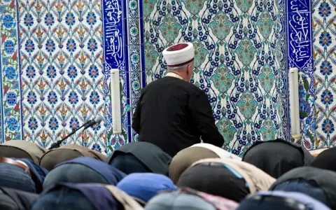 Männer in einer Moschee, die sich im Gebet verneigen. Der Imam trägt einen weißen und roten Hut. Die Fliesenwand ist mit islamischer Kalligrafie und Mustern verziert. Dies ist ein Ort, an dem der größte Sünde im Islam, Shirk, vermieden wird.