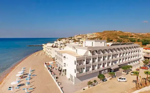 Wypoczynek na plaży w hotelu Island Maya Resort. Biały budynek hotelowy z balkonami, obok piaszczysta plaża z leżakami i parasolami, morze i wzgórza w tle.