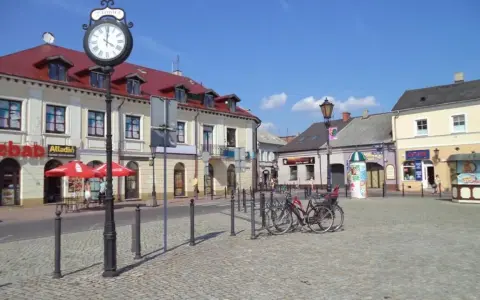 Słoneczny stary rynek łowicz z brukowanym placem, budynkami z czerwonymi dachami i zegarem na słupie.