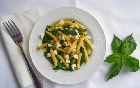 Pyszny makaron penne ze szpinakiem i twarogiem, udekorowany listkami bazylii. Obok leży widelec i biała serwetka.