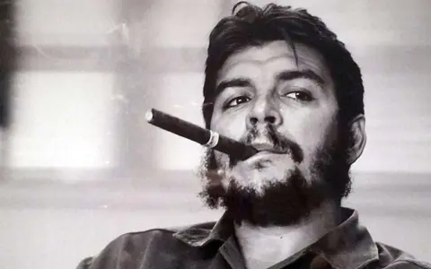 Portrait en noir et blanc de Che Guevara, cigare aux lèvres, regard intense. L'image évoque sa légende, même après sa mort.