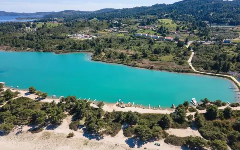 Turkusowa laguna na Kassandrze, otoczona zielonymi wzgórzami i lasem sosnowym. W oddali widać zabudowania i plażę z łodziami.
