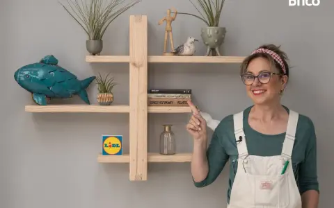 Una mujer sonriente muestra una estantería de madera, decorada con plantas, un pez azul y libros. Ideal para aprender como hacer una estantería de madera.