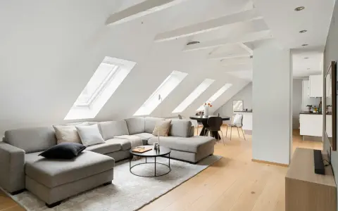 Soggiorno mansardato con divano angolare grigio, tavolino nero e travi a vista moderne dipinte di bianco che creano un'atmosfera luminosa.