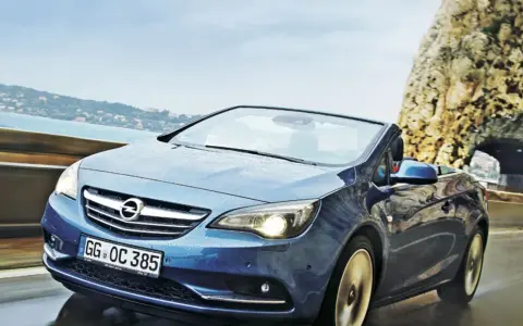 Niebieski Opel Cascada z opuszczonym dachem jedzie po mokrej drodze przy skalistym wybrzeżu.