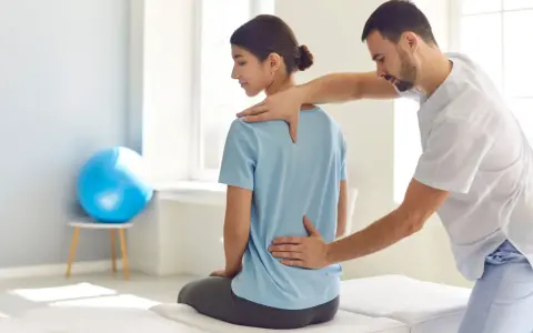 Fizjoterapeuta bada plecy pacjentki, sprawdzając, gdzie są lędźwie. W tle niebieska piłka do ćwiczeń.