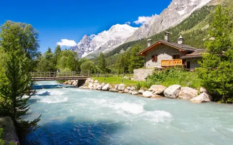 Val Ferret: un'incantevole casa in pietra alpina vicino a un fiume turchese, con un ponte e montagne innevate sullo sfondo.