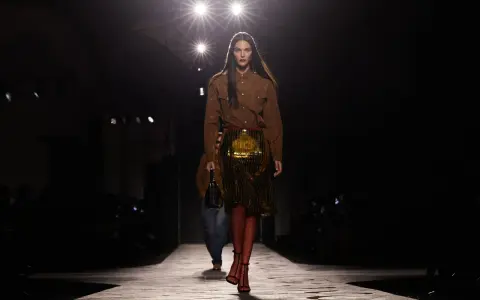 Modella su passerella, con gonna dorata e camicia marrone, un omaggio all'eleganza senza tempo di Prada, fondatore di un impero della moda.