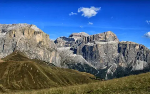 Vaste cime rocciose del gruppo Sella Dolomiti sotto un cielo azzurro con nuvole bianche. Prati verdi e sentieri serpeggiano sui pendii.