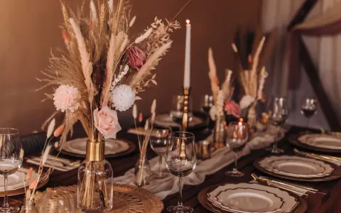 Stół weselny w stylu boho z bukietem z traw pampasowych, róż i świecami. Eleganckie nakrycie z złotymi talerzami i kieliszkami.