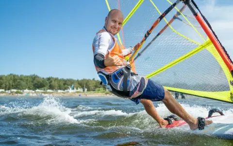 Mężczyzna na desce windsurfingowej, uśmiecha się i pokazuje kciuk w górę.