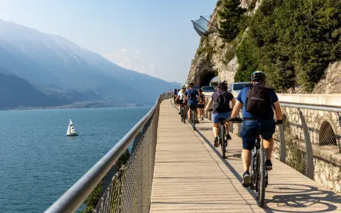 Grupa rowerzystów przemierza malownicze trasy rowerowe wokół jeziora, podziwiając góry i żaglówkę.
