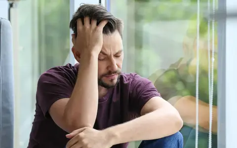 Mann sitzt mit geschlossenen Augen und der Hand am Kopf vor einem Fenster.