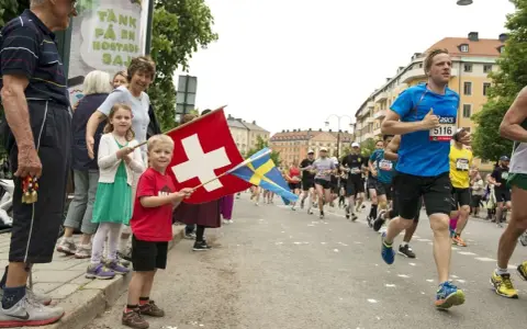 Barn viftar med schweizisk och svensk flagga vid ett lopp. Löpare på marathon distans passerar.