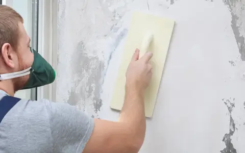 Mężczyzna w masce ochronnej nakłada tynk mozaikowy na ścianę za pomocą kielni.