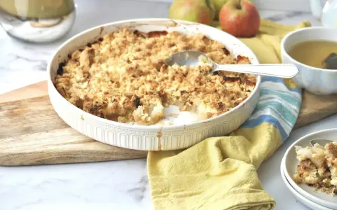 Ein goldbrauner Apfel-Crumble in einer Auflaufform, bereit zum Servieren. Daneben eine Tasse Tee und frische Äpfel.