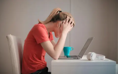 Kobieta z rękami na głowie, przy laptopie, z kubkiem i słuchawkami. Cierpi na uzależnienie od stresu, czuje się przytłoczona.
