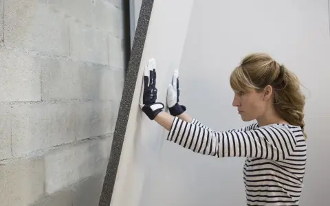 Une femme en gants manipule une plaque de doublage polystyrène expansé contre un mur de parpaings.