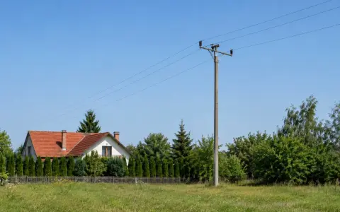 Słup na działce, obok domu z czerwoną dachówką i żywopłotem. W tle zielone drzewa i błękitne niebo.