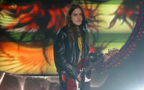 Zalewski w Idolu, z gitarą i w skórzanej kurtce, na tle kolorowej scenografii.