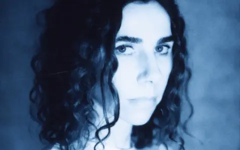 PJ Harvey, z kręconymi włosami, patrzy intensywnie. Jej twarz, w odcieniach niebieskiego, zdaje się opowiadać historię z "I Inside the Old I Dying".