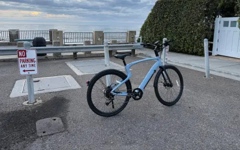 A light blue electric city bike is parked on asphalt near a "No Parking" sign, with the ocean and cloudy sky in the background.