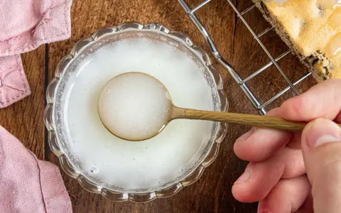Ein Löffel mit einer weißen, schaumigen Masse, die zeigt, was ist Staubzucker, in einer Glasschale. Daneben ein Stück Kuchen.