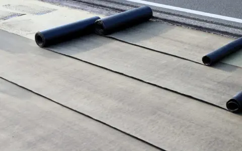 Instalación de tela asfáltica en rollo sobre una superficie. Se aprecian varias capas desplegadas y rollos listos para continuar.