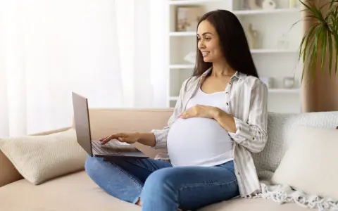 Kobieta w ciąży na kanapie szuka w internecie najdokładniejszy termin porodu, uśmiecha się.