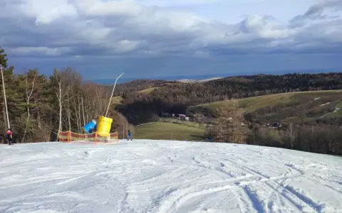 Śnieżny stok narciarski z armatkami śnieżnymi i widokiem na pagórkowaty krajobraz.