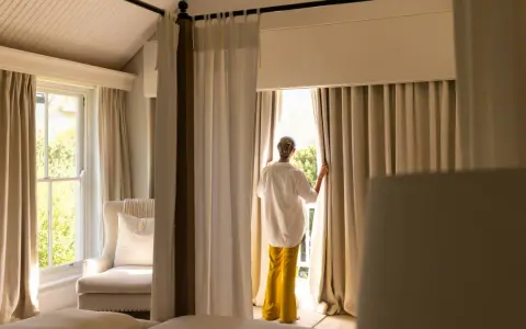 Una mujer abre cortinas para dormitorios, dejando entrar la luz. Se ven camas y un sillón junto a la ventana.