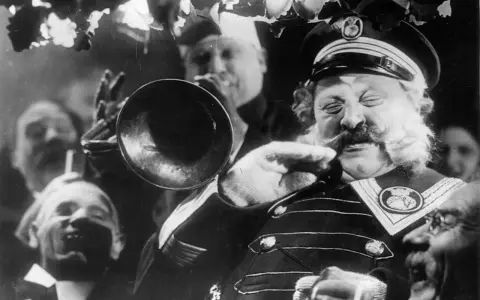 Un uomo con baffi e barba folta suona una tromba, circondato da volti sorridenti. Un'atmosfera da kammerspiel, un dramma intimo e intenso.
