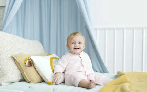 Uśmiechnięte, 4-miesięczne dziecko siada na łóżku w otoczeniu niebieskiego baldachimu i kolorowych poduszek.