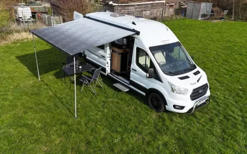 Vit husbil, Karmann Dexter, med utdraget markis och campingmöbler på gräs.