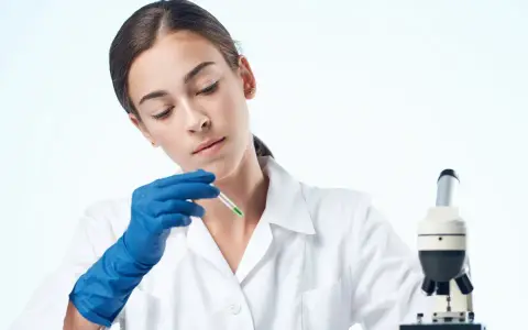 Giovane tecnologo alimentare in camice bianco, con guanti blu, analizza un campione con un contagocce accanto a un microscopio.