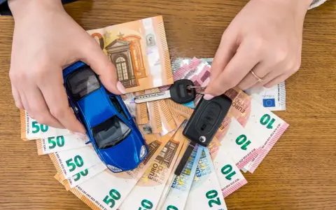 Manos sostienen un coche azul de juguete, llaves y billetes de euro. El coche no arranca, quizás por falta de dinero.