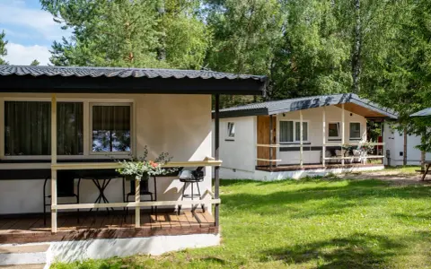 Dwa białe domki kempingowe w lesie, z werandami i meblami ogrodowymi. Idealne miejsce dla każdego **camping wagabunda**.