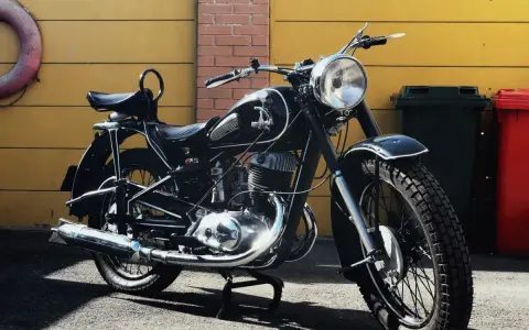 Klasyczny czarny motocykl, model iż 49, lśni w słońcu. Obok żółta brama i ceglana ściana.