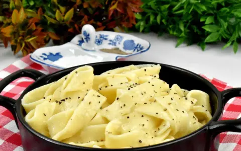 Pappardelle w kremowym sosie alfredo, posypane świeżo mielonym pieprzem, podane w czarnym naczyniu na czerwono-białej kratce.