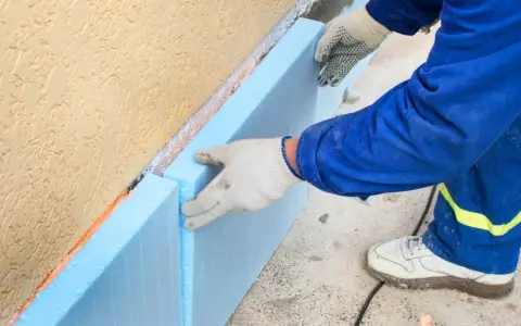 Ręce w rękawiczkach układają niebieski styropian przy ocieplanej ścianie fundamentowej.