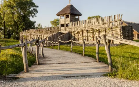 Drewniana kładka prowadzi do grodziska, jednego z historycznych miejsc w Polsce. Widać palisadę i wieżę obronną.