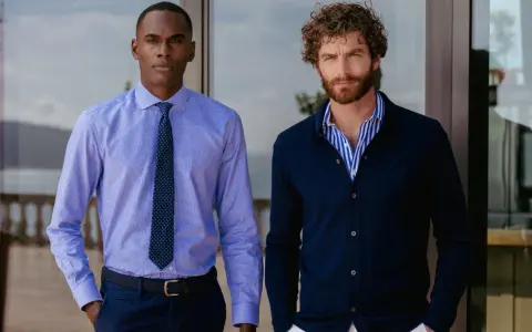 Dos hombres posan. Uno lleva una camisa de vestir azul con corbata, la camisa por dentro. El otro, una camisa de rayas azules y blancas con cárdigan azul oscuro, la camisa por fuera.