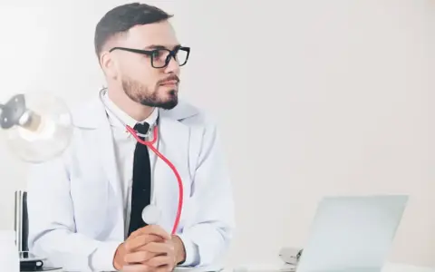 Lekarz medycyny pracy w okularach, z czerwonym stetoskopem, siedzi przy biurku z laptopem. Jakie badania wykonuje?